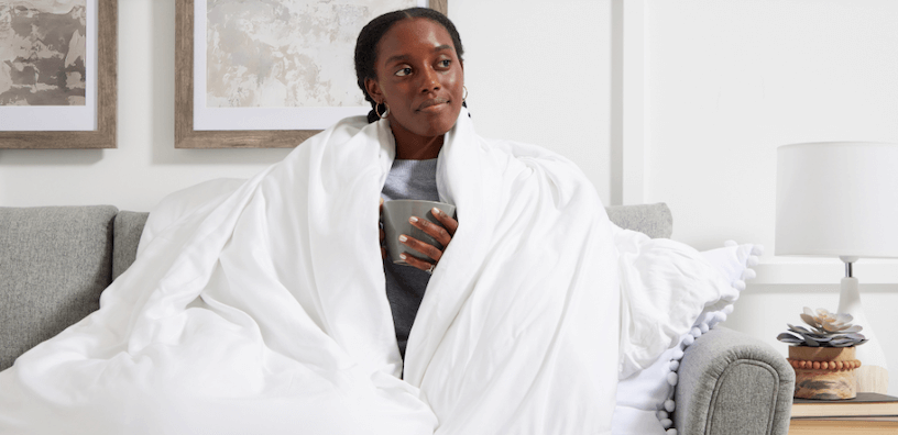 Une femme assise sur un canapé confortable, enveloppée dans une douce couverture blanche, tenant une tasse à la main, et l’air détendu, créant une atmosphère calme et réconfortante.